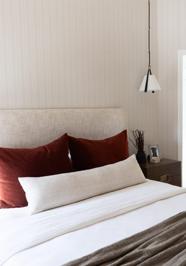 A bed with rust-colored pillows, a long white cushion, beige headboard, brown throw, and bedside table with lamp and vase— Living with Lolo.