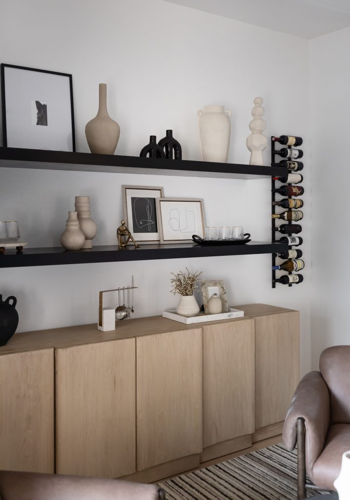 Modern shelving displays ceramics, framed art, glassware, and a wine rack above a light wood cabinet and neutral chairs. — Living with Lolo.