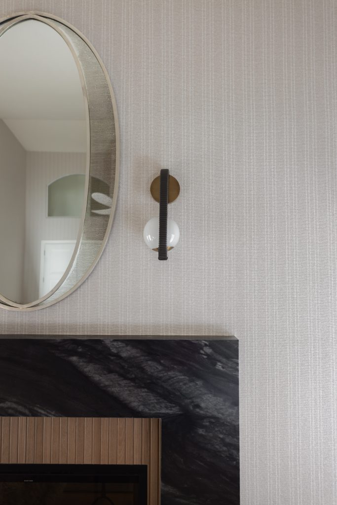 An oval mirror, a wall sconce with a white bulb, and a black marble mantelpiece on light striped wallpaper—Living with Lolo.