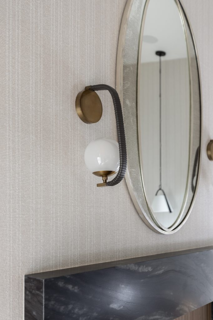 A wall-mounted sconce with a round bulb is beside a large oval mirror above a black marble surface. A ceiling light fixture appears in the mirror—Living with Lolo.