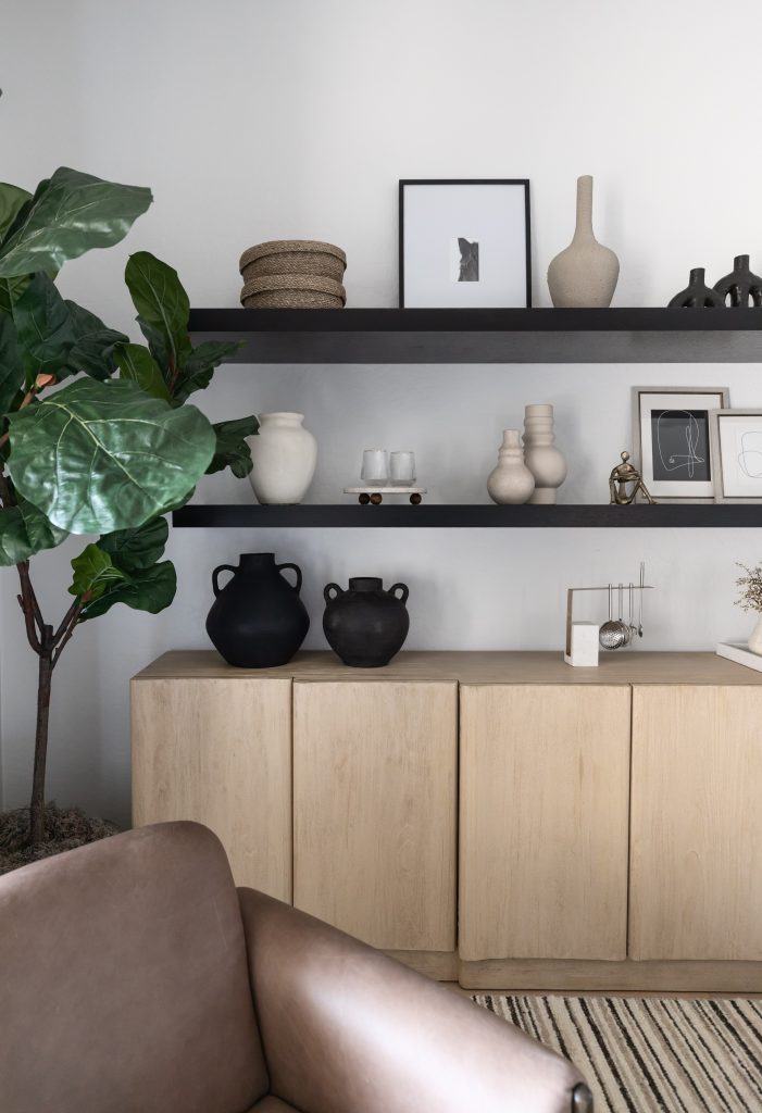 A tan armchair sits beside a light wood cabinet, black shelves with vases, framed art, and a large green potted plant— Living with Lolo.