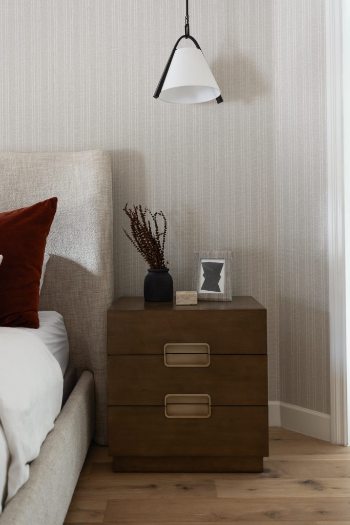 A wooden nightstand with two drawers sits next to a bed, with a small vase, framed abstract art, and a pendant light above. — Living with Lolo.