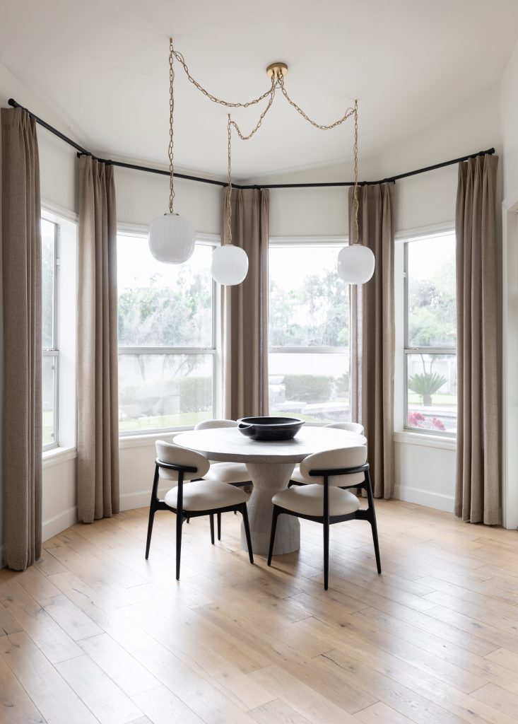 A dining nook with a round white table, four black and white chairs, beige curtains, light wood floors, and three pendant lights. — Living with Lolo.