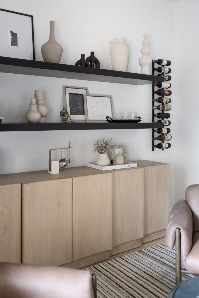 A living room corner with a light wood cabinet, black shelves, ceramics, framed art, wine rack, and a striped rug—Living with Lolo.