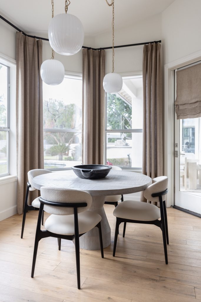 A round dining table with four chairs is set in a bright room featuring large windows, beige curtains, wood floors, and three white pendant lights overhead — Living with Lolo.