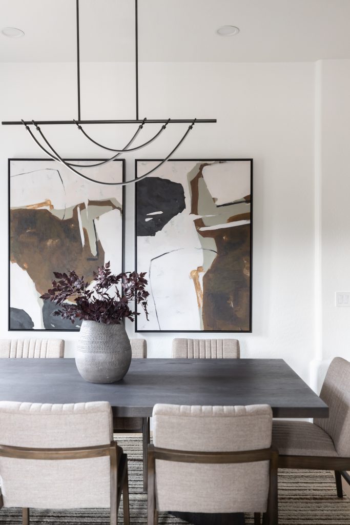 A dark rectangular dining table with beige upholstered chairs, a gray vase of dark foliage, and abstract art on a white wall. — Living with Lolo.