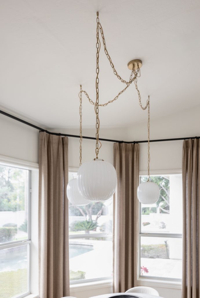 Three white globe pendant lights with gold chains hang from the ceiling, near beige curtains and large windows. — Living with Lolo.