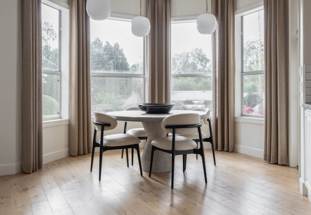 A round dining table with four chairs is placed in a bay window area with beige curtains, wooden floors, and three pendant lights— Living with Lolo.