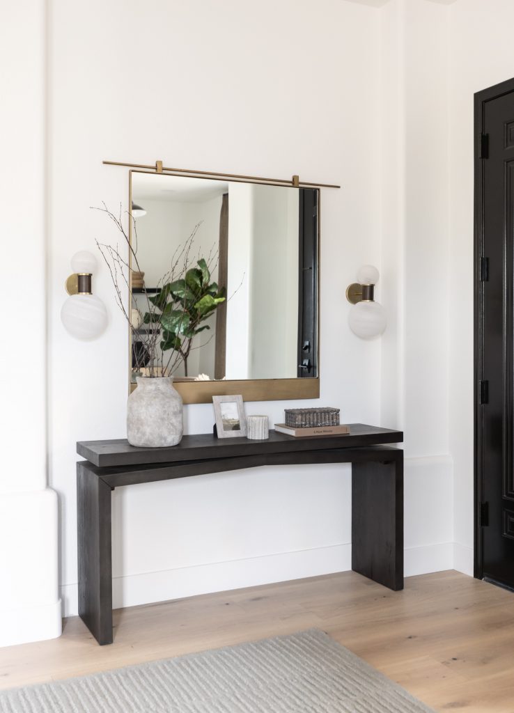 A modern console table holds a vase of branches, a framed photo, and books beneath a large mirror with wall sconces—Living with Lolo.