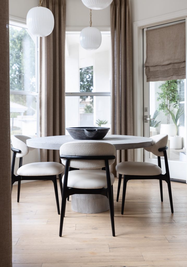 A round dining table with four chairs sits below white pendant lights, near large windows with beige curtains and a black bowl centerpiece— Living with Lolo.