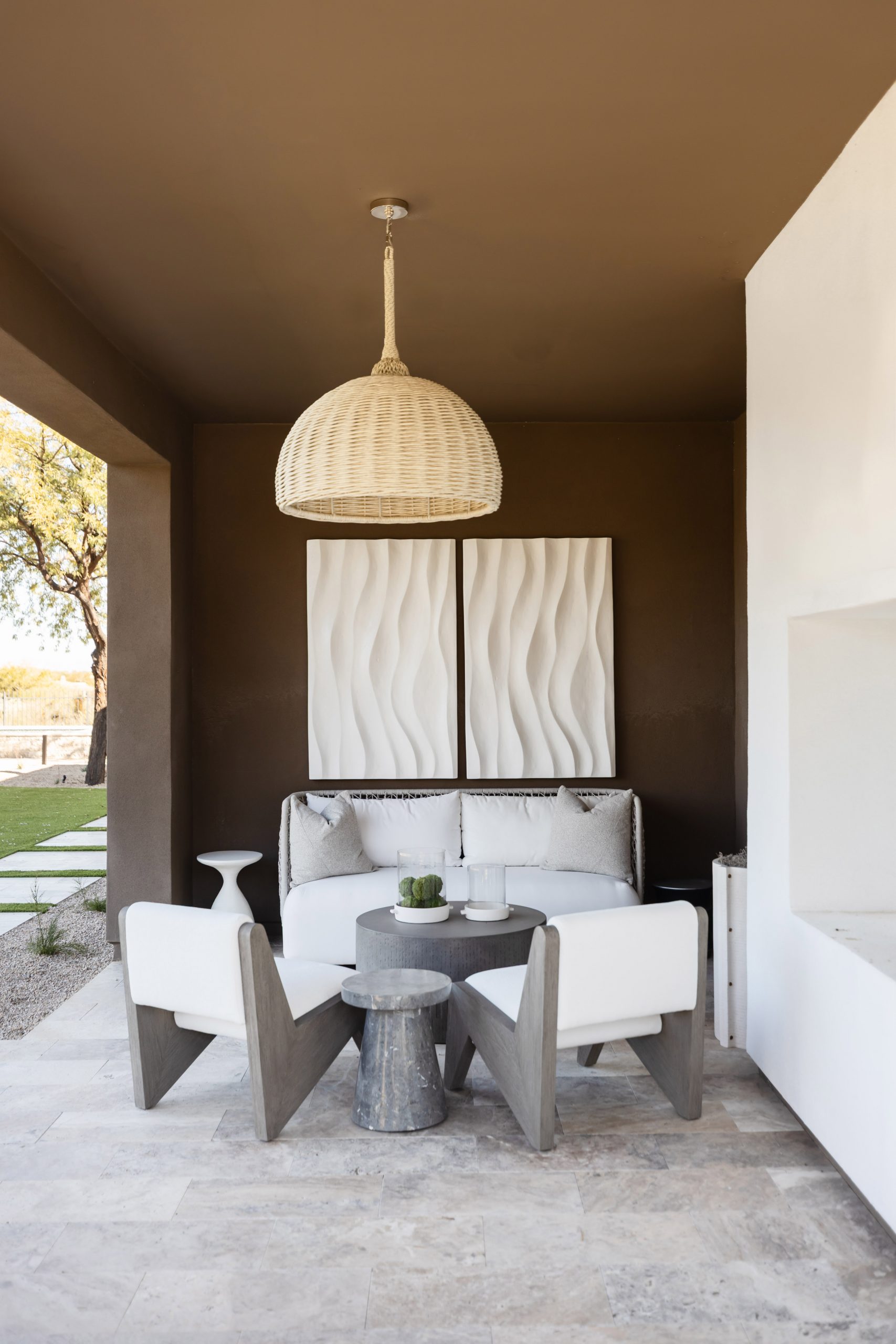 White cushioned chairs surround a round table on a patio with textured wall art and a woven pendant light.— Living with Lolo.