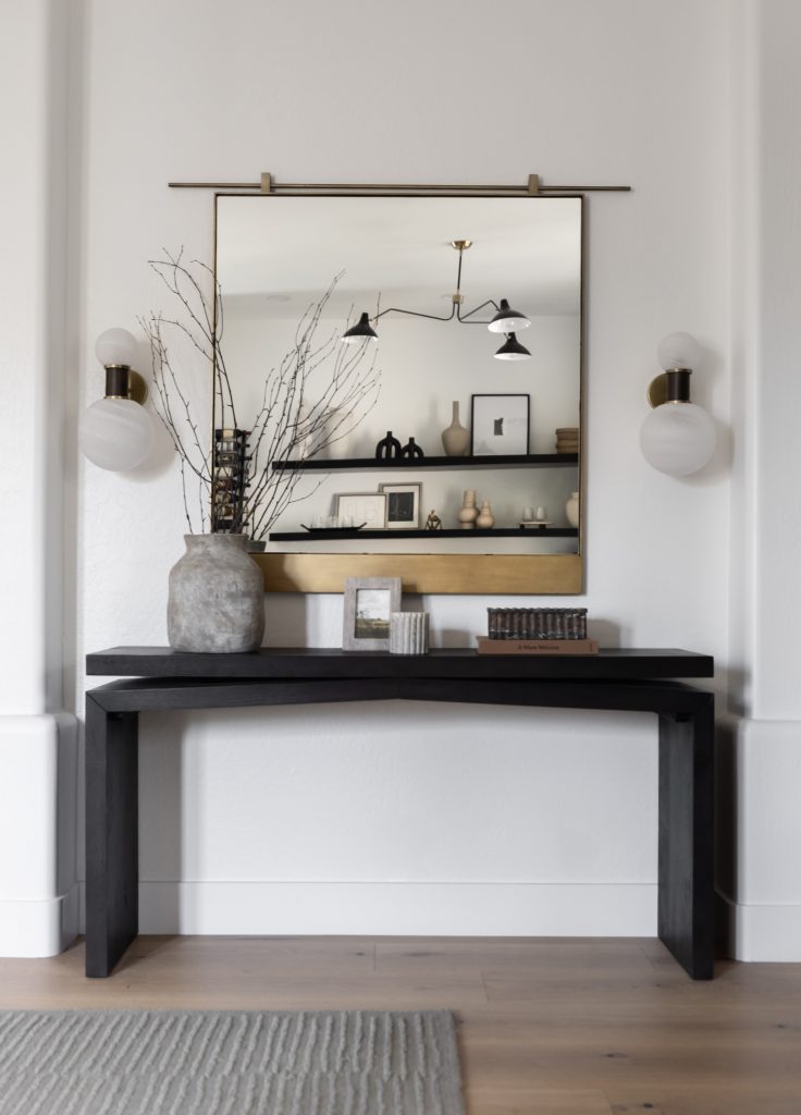 A black console table sits below a large square mirror, with wall sconces, a vase with branches, framed photos, and books. — Living with Lolo.
