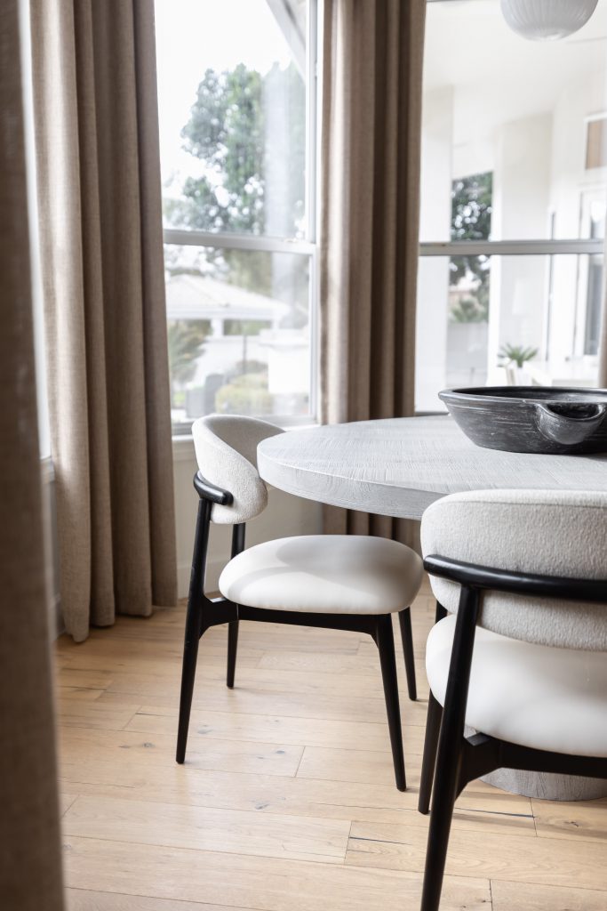 Round dining table with two white and black chairs, beige curtains on large windows, and light wood floors. — Living with Lolo.