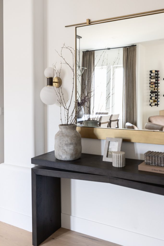 A black console table with a stone vase, branches, picture frames, and books sits below a large mirror in a modern living room — Living with Lolo.