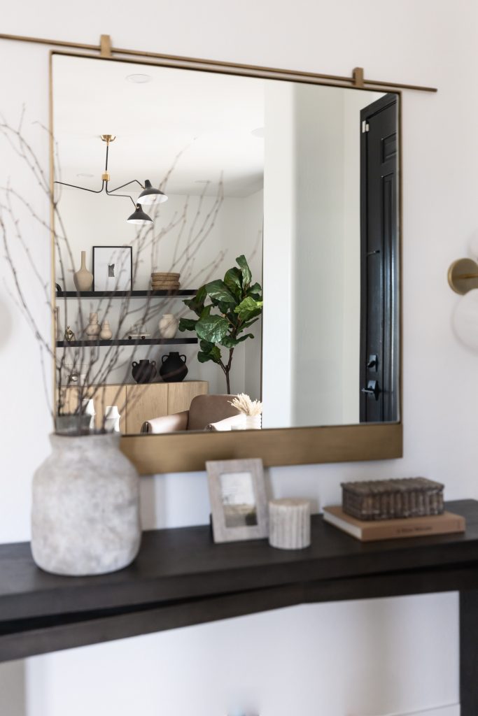A rectangular mirror above a dark console table reflects shelves, wall decor, and greenery in a modern living room. — Living with Lolo.