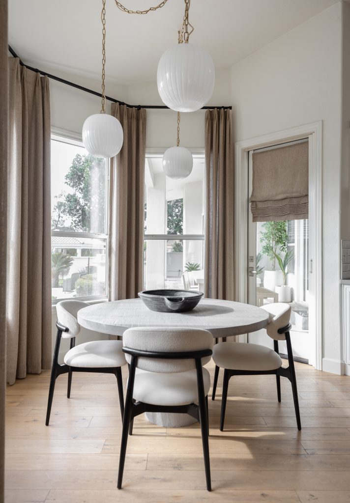 A dining area with a round table, five cushioned chairs, black bowl centerpiece, large windows, beige curtains, and white pendant lights — Living with Lolo.
