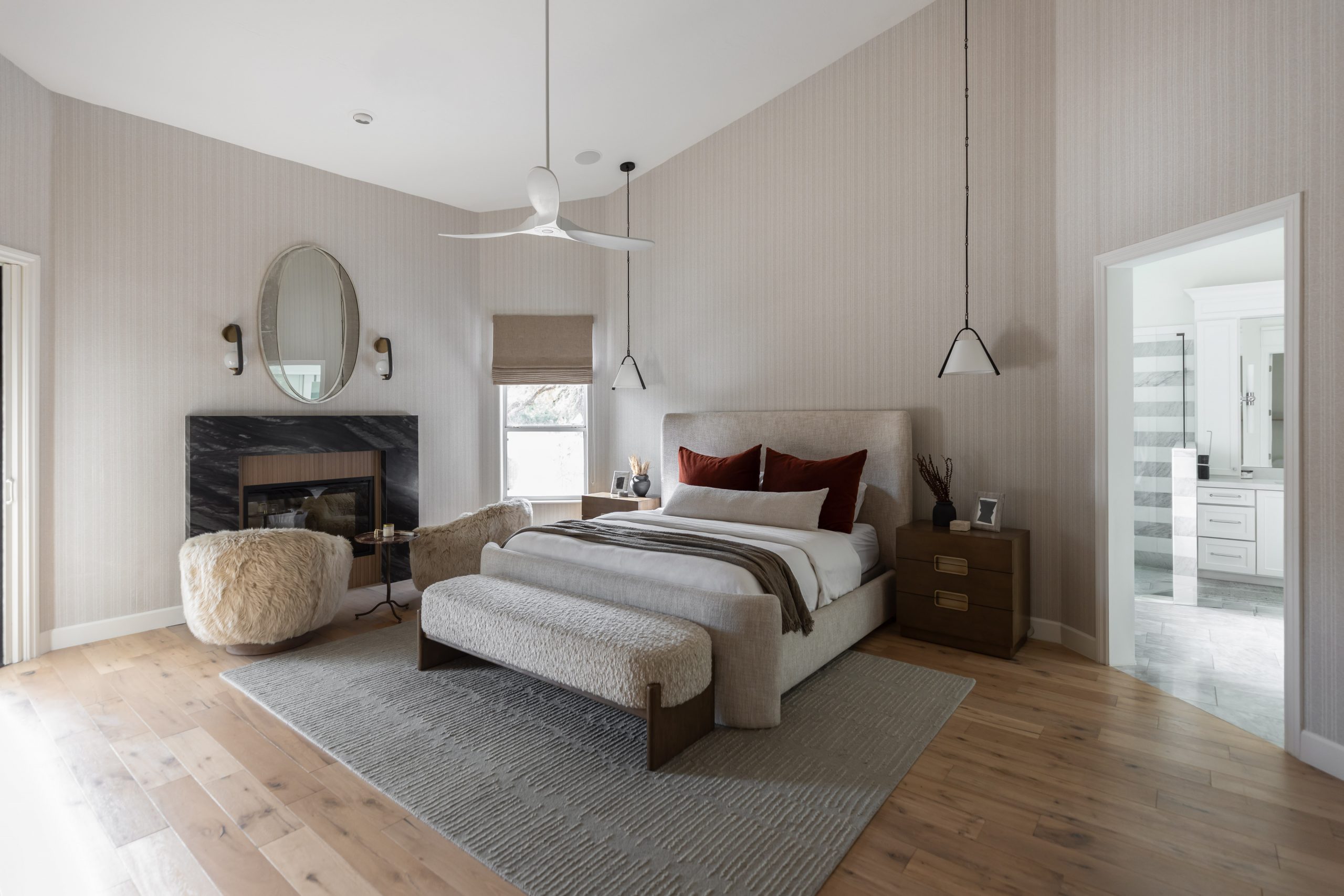 A modern bedroom includes a beige bed with colored pillows, a fireplace, an armchair, a ceiling fan, and an attached bathroom with white cabinets. — Living with Lolo.