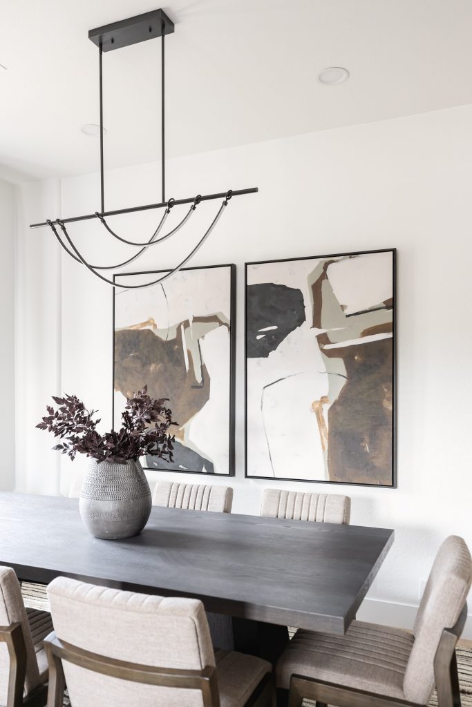 A dining room with a dark rectangular table, six beige chairs, a gray vase with dark foliage, abstract wall art, and a black metal light fixture — Living with Lolo.