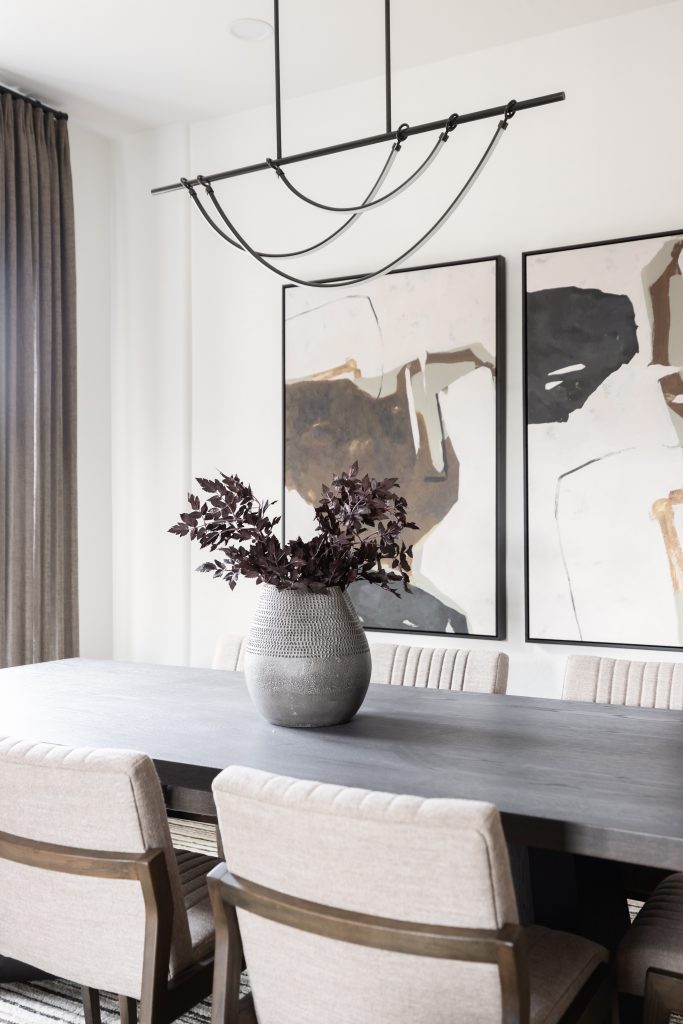A dining room with a dark wooden table, six upholstered chairs, a gray vase with dark foliage, abstract wall art, and a black metal chandelier — Living with Lolo.