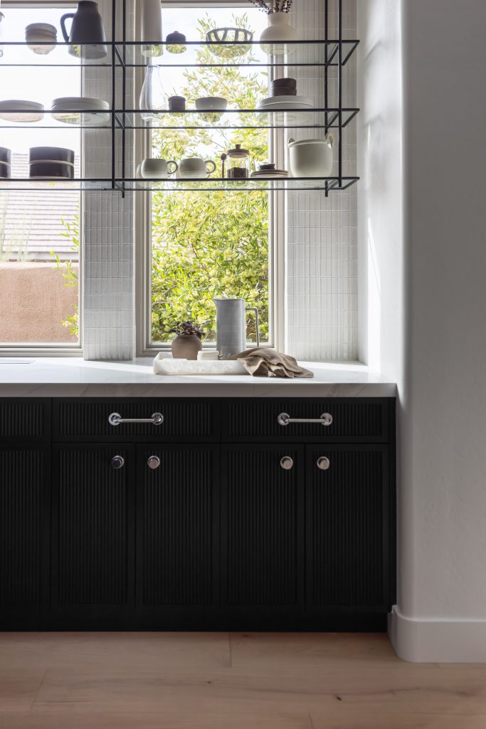 Modern kitchen featuring black lower cabinets, a white countertop, open shelves with dishes, and greenery visible through the window—Living with Lolo.