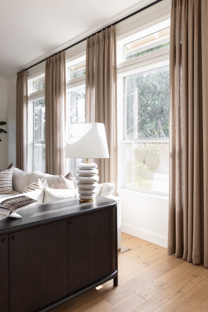 A living room features tall windows with beige curtains, a white lamp on a dark console, a light sofa, and wood flooring—Living with Lolo.