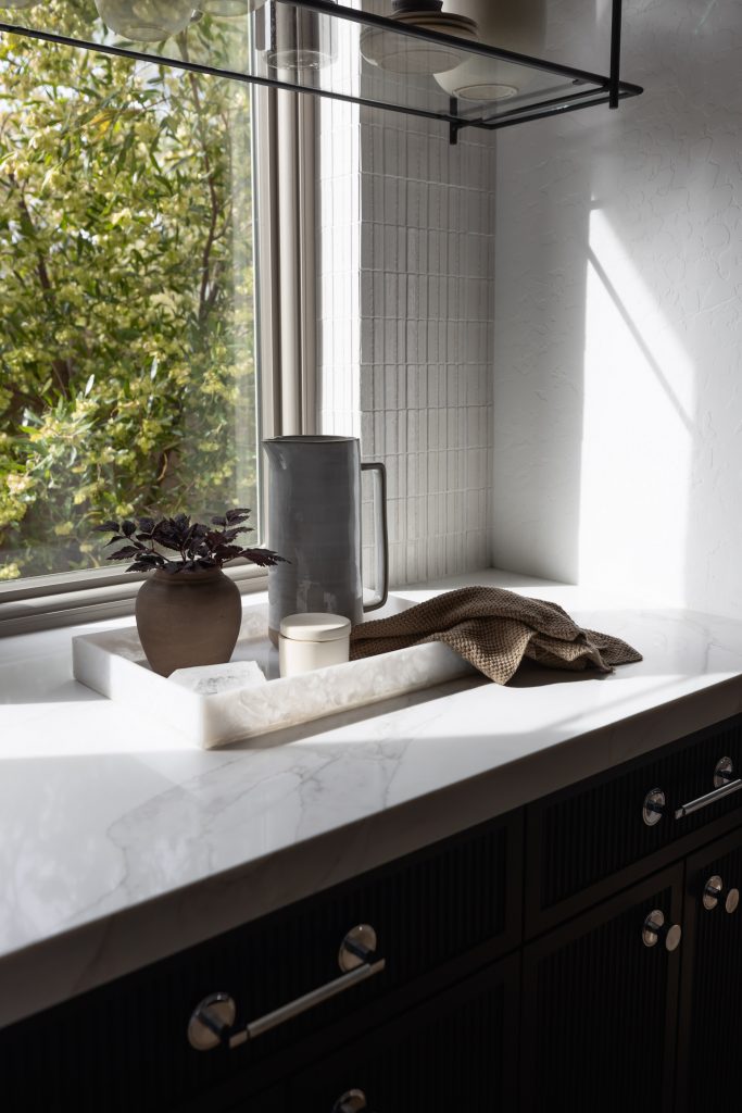 A marble tray holding a vase, cup, pitcher, and folded towel is on a white countertop beside a window with a view of greenery — Living with Lolo.