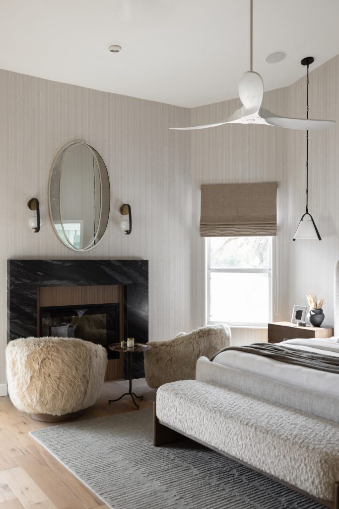 Modern bedroom featuring a marble fireplace, sheepskin chairs, bed, large oval mirror, wall sconces, window shade, and ceiling fan—Living with Lolo.