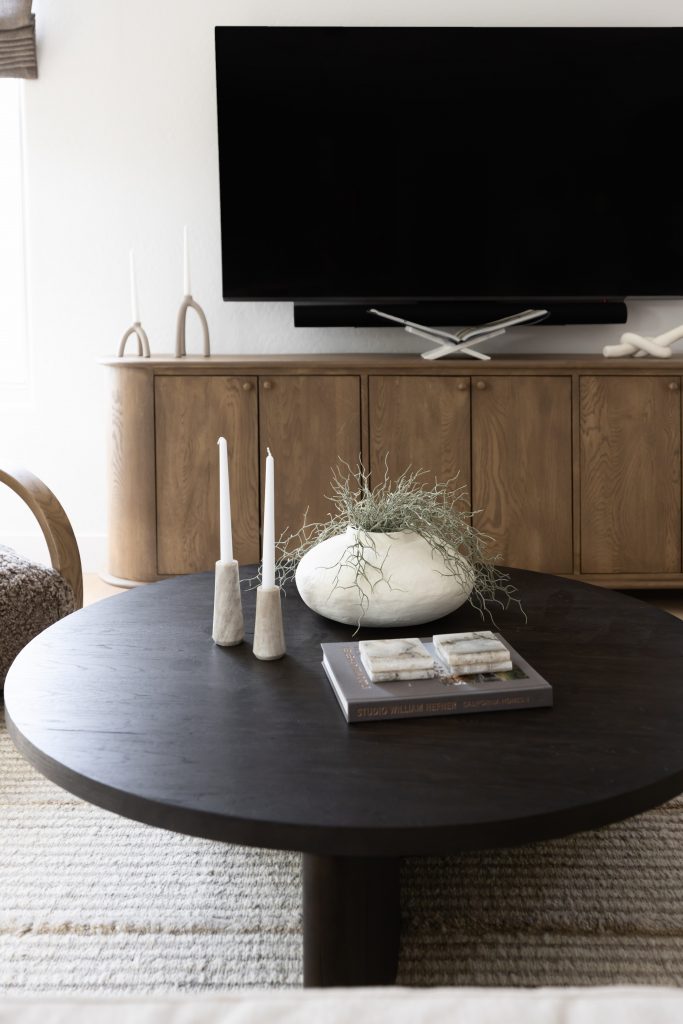 A round black coffee table with candles, a vase of greenery, and a book is in front of a wooden media console and wall-mounted TV — Living with Lolo.
