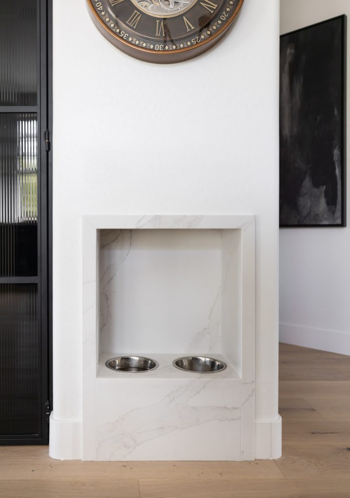 A built-in pet feeding station with two stainless steel bowls is set into a white marble alcove beneath a wall clock — Living with Lolo.