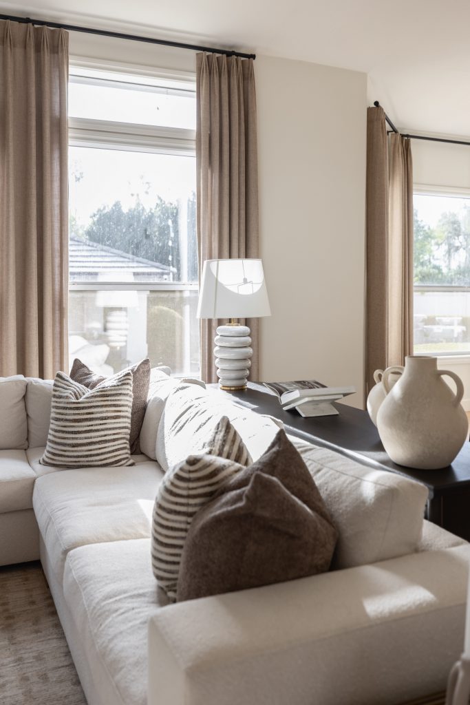 A bright living room features a white sectional sofa, throw pillows, a black console table, ceramic vases, and large windows—Living with Lolo.
