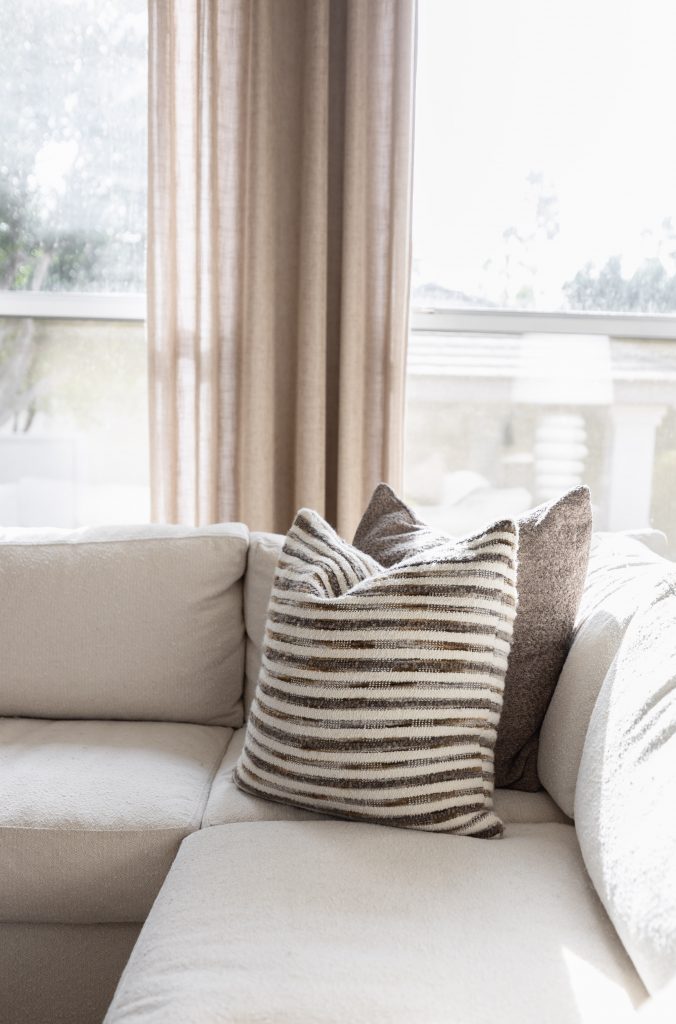 A beige sectional sofa with a striped throw pillow and a solid pillow sits by a window with sheer curtains in daylight — Living with Lolo.