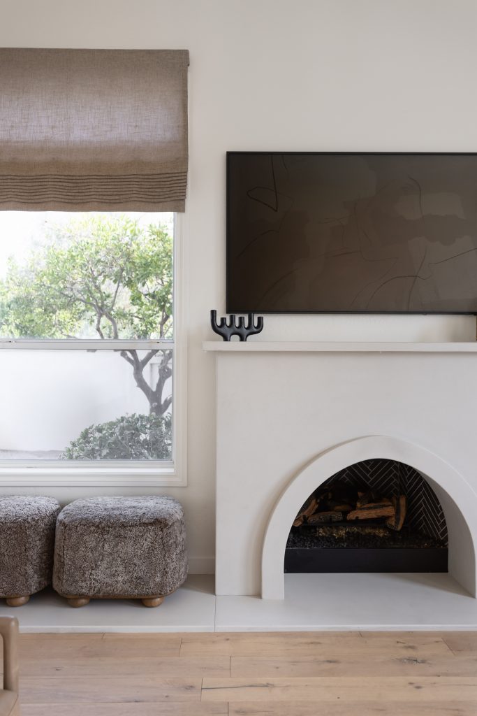 A white arched fireplace with a TV above, two textured ottomans, and a window with a beige shade are shown—Living with Lolo.