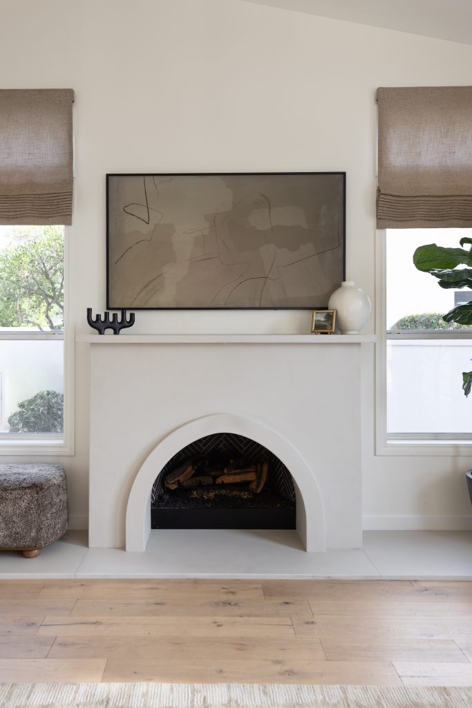 A modern white fireplace features an abstract painting, black candleholder, white vase, and framed photo between two windows with beige Roman shades— Living with Lolo.