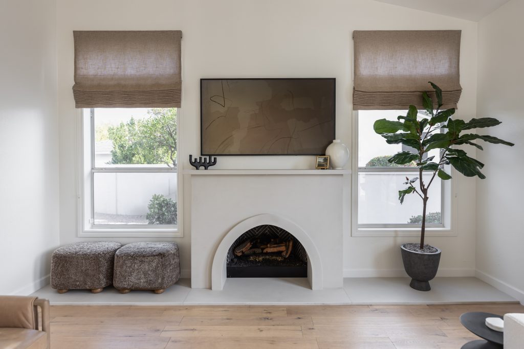 A modern living room shows a fireplace with a TV above, two ottomans, a potted plant, and two windows with beige Roman shades.— Living with Lolo.