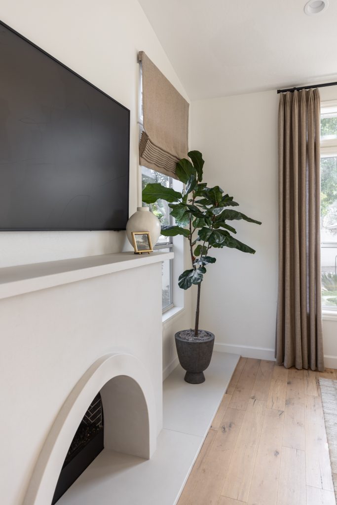 A wall-mounted TV above a white fireplace, a potted plant, beige curtains, and wooden floors are visible—Living with Lolo.