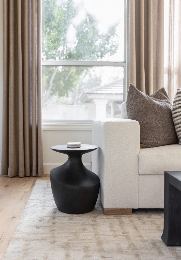 A white sofa with gray and striped pillows sits next to a black sculptural side table and beige curtains by a large window. — Living with Lolo.