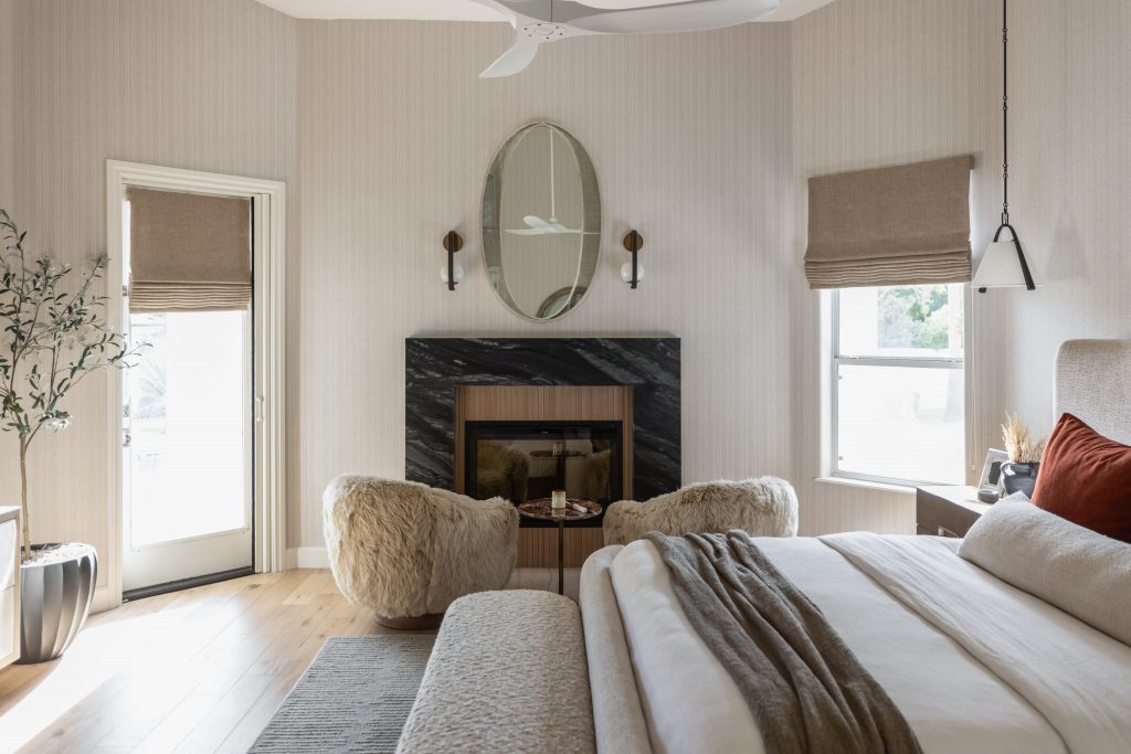 Modern bedroom with neutral colors, a fireplace, two fuzzy chairs, an oval mirror, and beige Roman shades on the door and window — Living with Lolo.