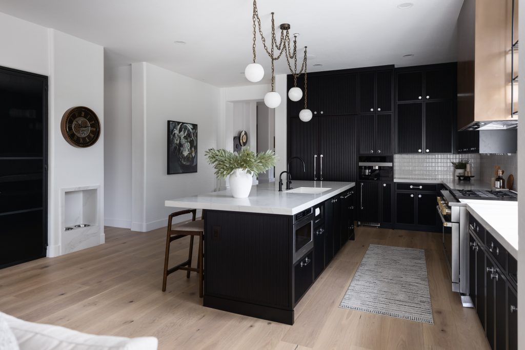 Modern kitchen with black cabinets, white countertops, a central island with two stools, pendant lights, and light wood flooring—Living with Lolo.