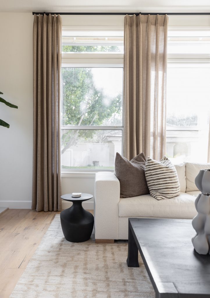 A modern living room with beige curtains, a white sofa, neutral pillows, black tables, a light rug, and large windows. — Living with Lolo.