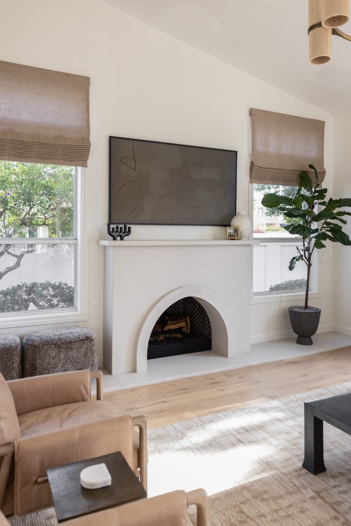 A living room with a curved white fireplace, large TV above, beige armchairs, potted plant, and windows with brown Roman shades. — Living with Lolo.