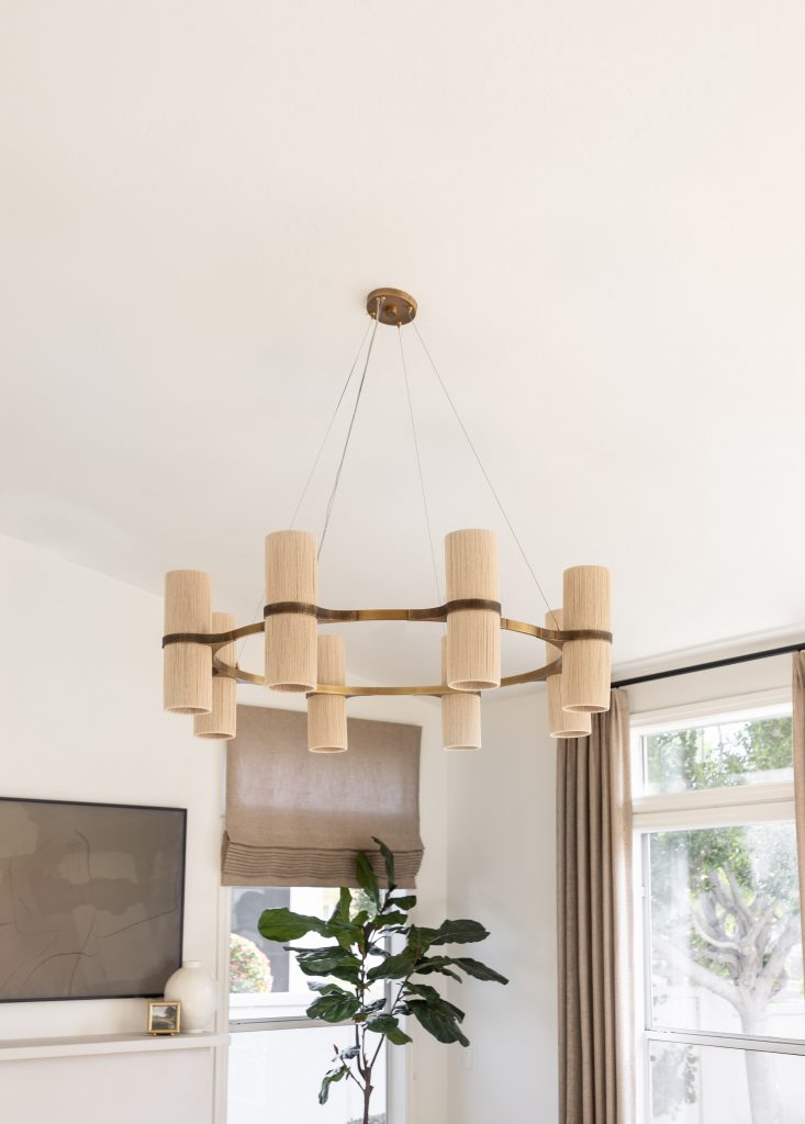 A modern chandelier with cylindrical beige shades hangs in a living room with large windows, curtains, a plant, and a wall-mounted TV. — Living with Lolo.