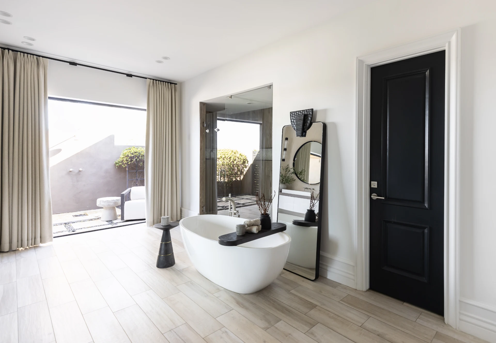 Freestanding tub, black door, large mirror, side tables, wood-look floor, and sliding glass doors to an outdoor patio—Living with Lolo.
