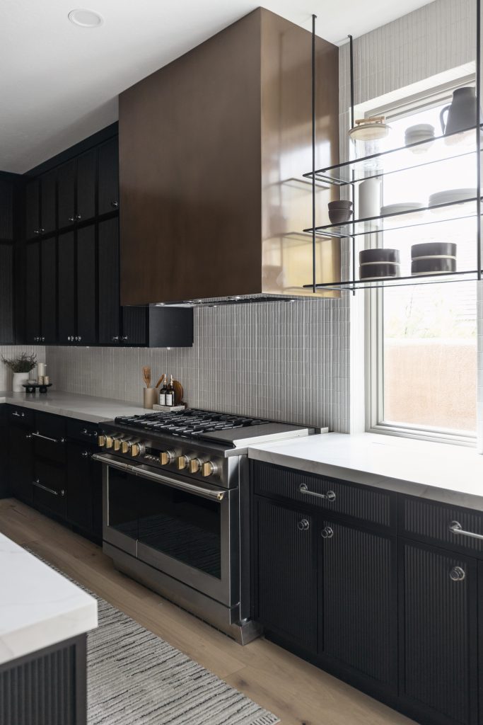 Modern kitchen with black cabinets, stainless steel stove, bronze range hood, open shelves, and a large window.— Living with Lolo.