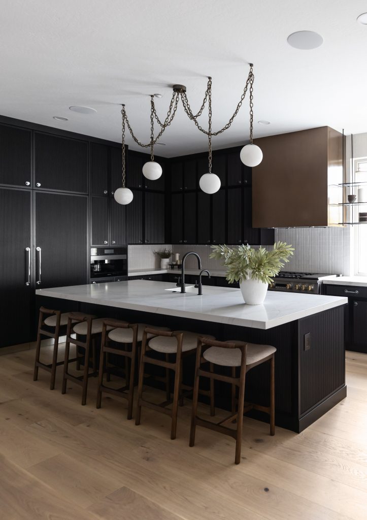 Modern kitchen with black cabinets, a marble island countertop, four wooden bar stools, a white potted plant, and globe lights—Living with Lolo.