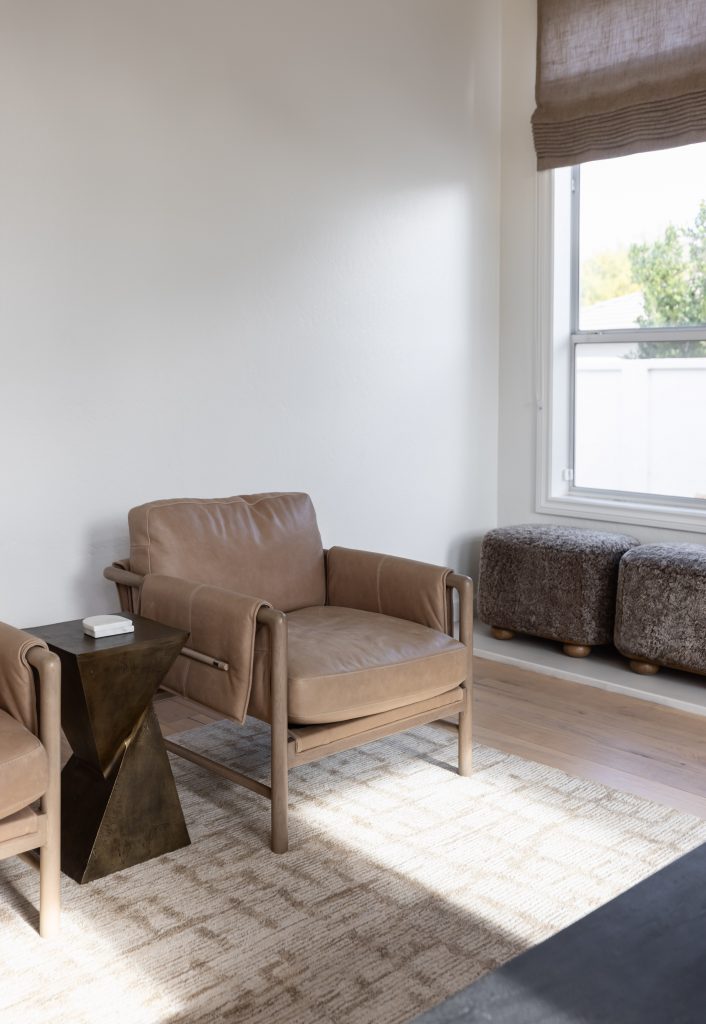 A living room with a tan leather armchair, dark geometric side table, patterned rug, two fuzzy ottomans, and a sunlit window—Living with Lolo.