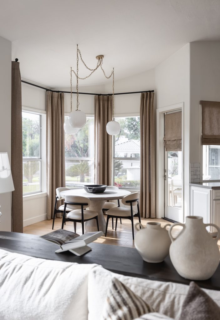 A round table with four chairs sits near large windows with beige curtains, pendant lights, and neutral-toned decor. — Living with Lolo.