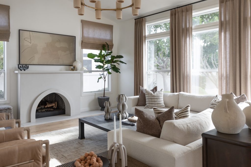 Bright living room with neutral furniture, a white fireplace, large windows with sheer curtains, potted plant, and abstract art—Living with Lolo.