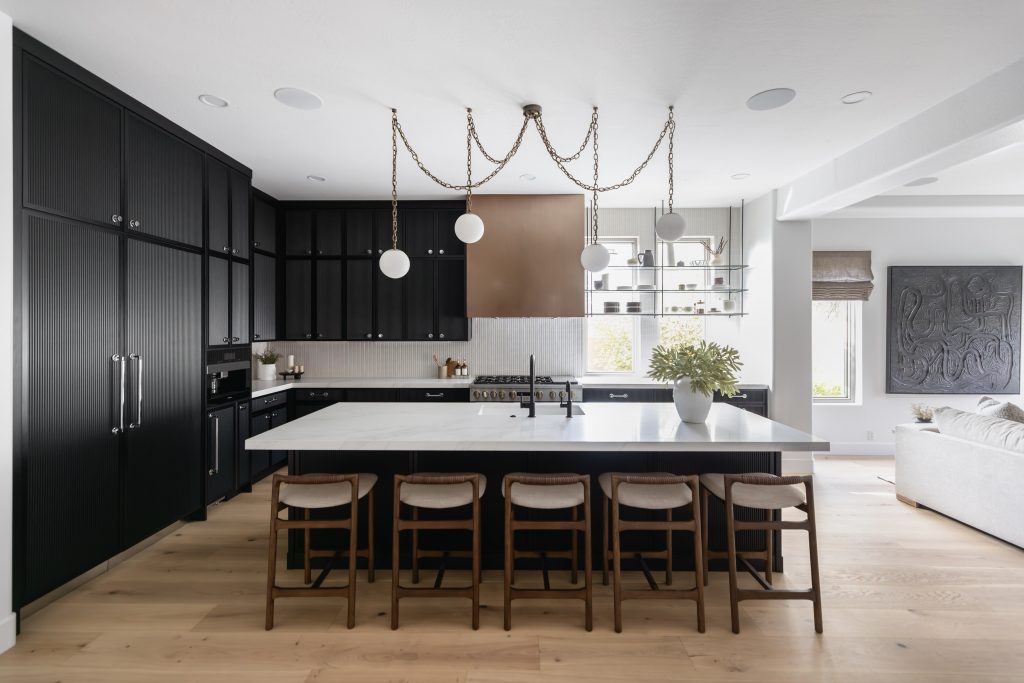 Modern kitchen with black cabinets, a white island, five wooden stools, pendant lights, open shelving, and light wood flooring—Living with Lolo.