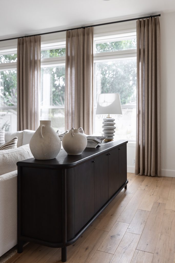 A modern living room with a dark sideboard, ceramic vases, stacked books, a table lamp, light curtains, and large windows. — Living with Lolo.