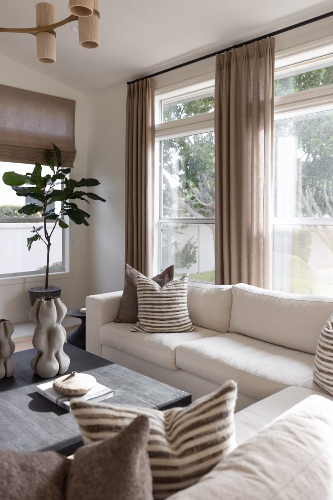 Beige sofas, striped throw pillows, a black coffee table, large windows with brown curtains, and a potted plant. — Living with Lolo.